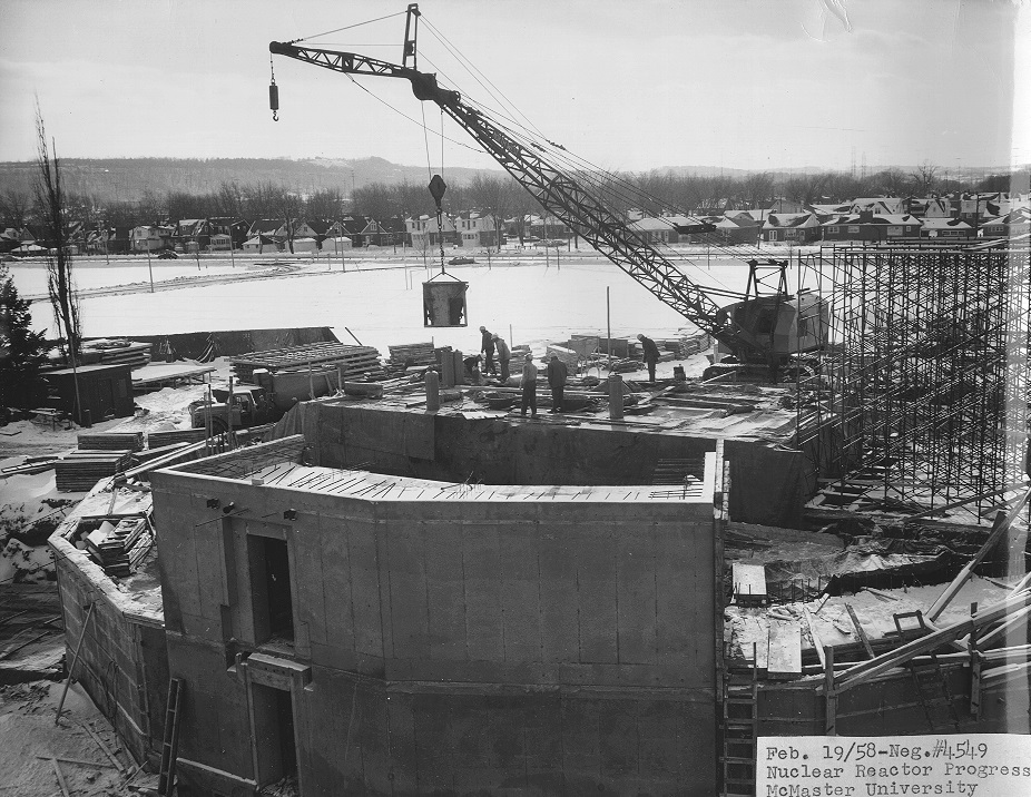 Construction workers building the McMaster Nuclear Reactor. Crane lifts materials in background.