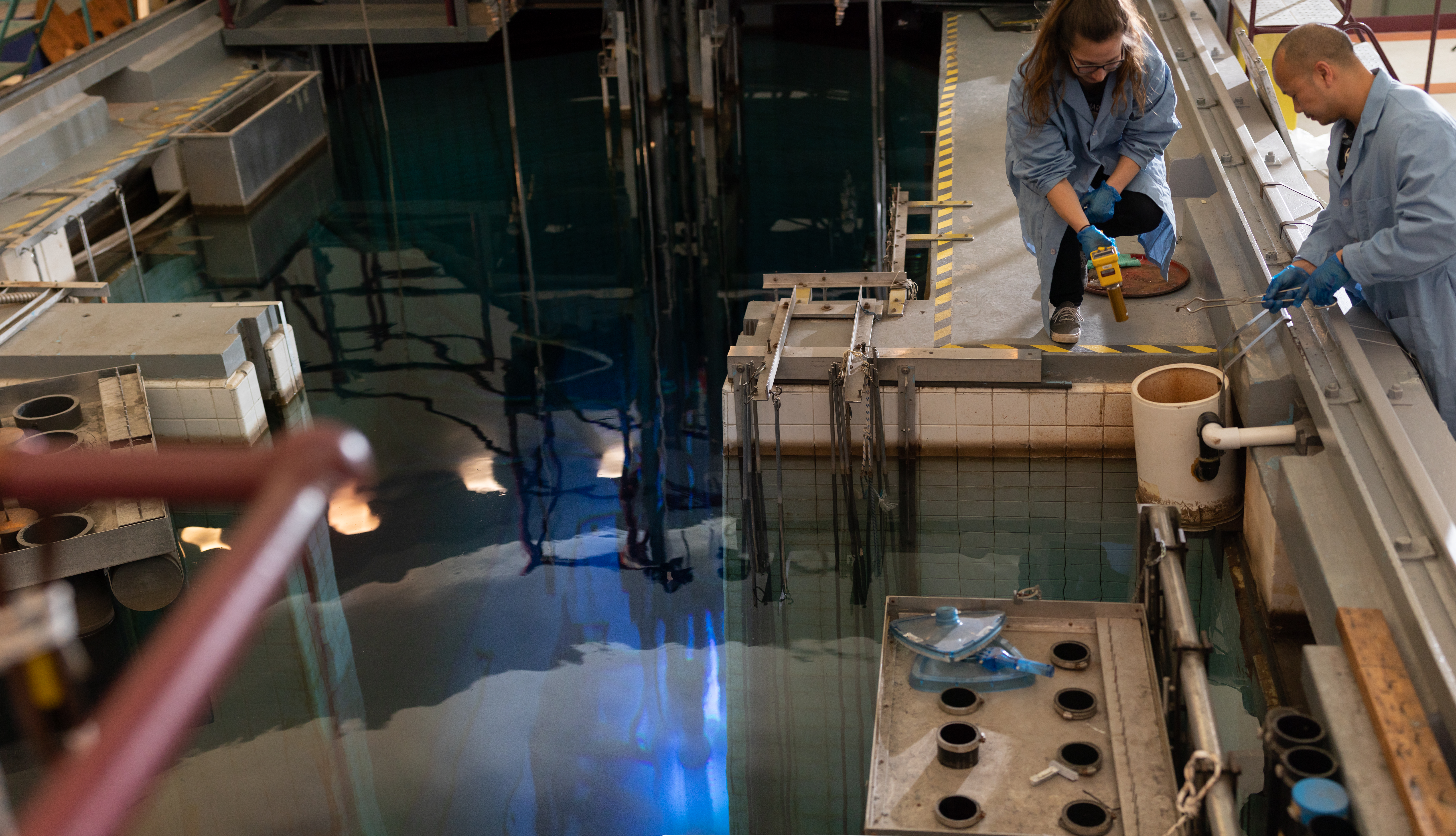 Two technicians in labcoats handle tools next to an open-pool nuclear reactor.